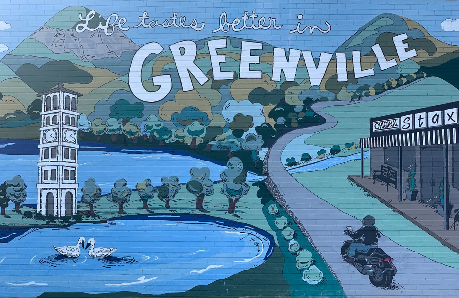 Wall mural of "Life tastes better in Greenville" with hills, a clocktower, a person on a motorcycle, and the Original Stax building